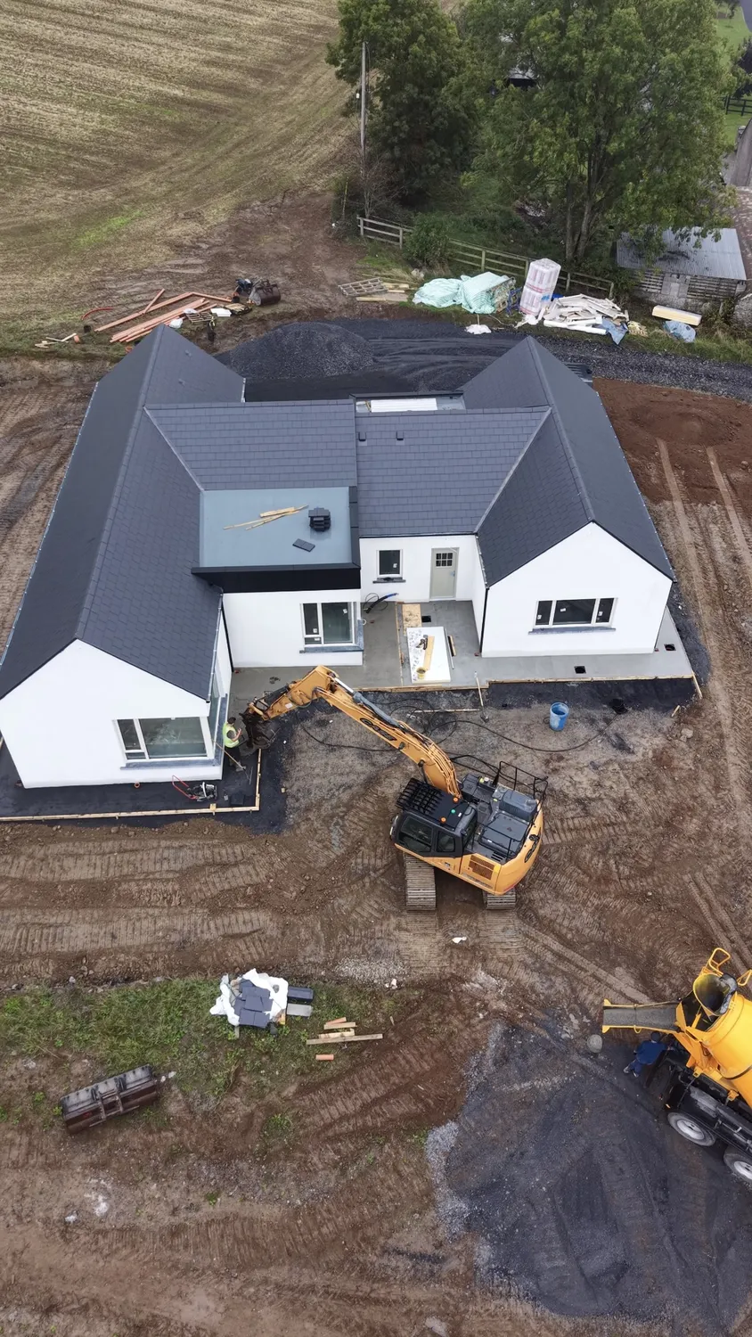 Worker smoothing concrete while an excavator works beside a house