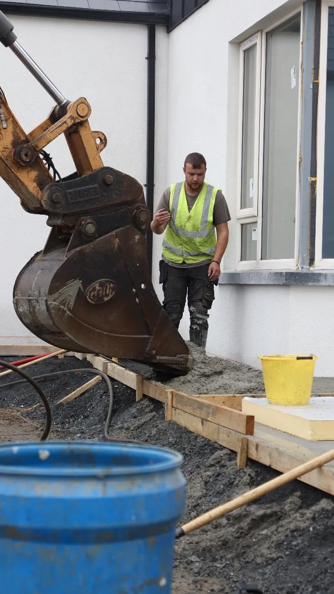 Worker and excavator at a fresh concrete path beside a house